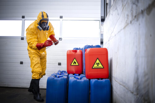 Worker in yellow protective suit, gas mask and gloves checking inventory of chemicals inside production plant storage room.